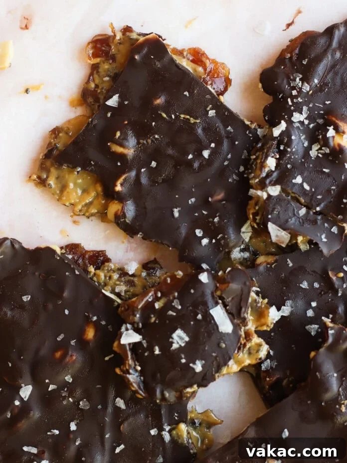 Snickers Inspired Peanut Butter Date Bark 9 Chocolate-covered brittle pieces topped with flaky sea salt, revealing layers of nuts and a caramel-like filling on a light surface.