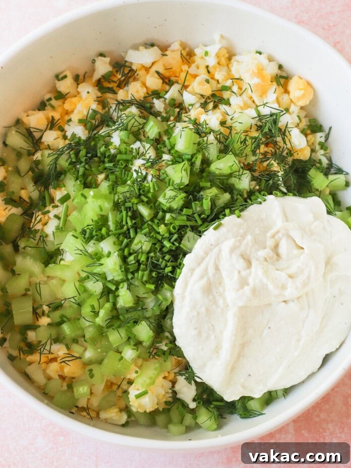 Creamy High Protein Cottage Cheese Egg Salad No Mayo 7 A white bowl filled with chopped hard-boiled eggs, celery, fresh dill, chives, and a large dollop of creamy mayonnaise or dressing, all ready to be mixed together.