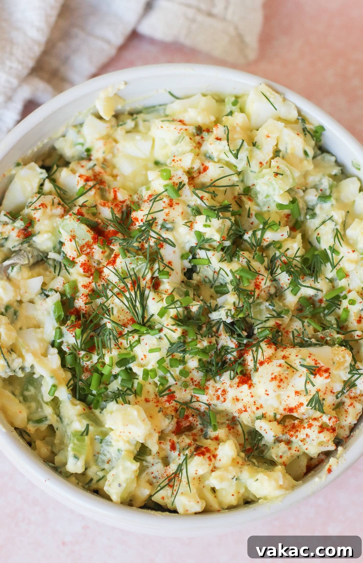 Creamy High Protein Cottage Cheese Egg Salad No Mayo 8 A bowl of creamy egg salad garnished with chopped chives, fresh dill, and a sprinkle of paprika, set on a light pink surface with a textured cloth in the background.