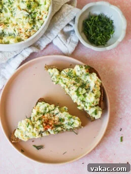 Two slices of bread topped with creamy egg salad and herbs are on a pink plate. Nearby are a bowl of egg salad, a small dish of chopped chives, and a light-colored cloth napkin on a pink surface.