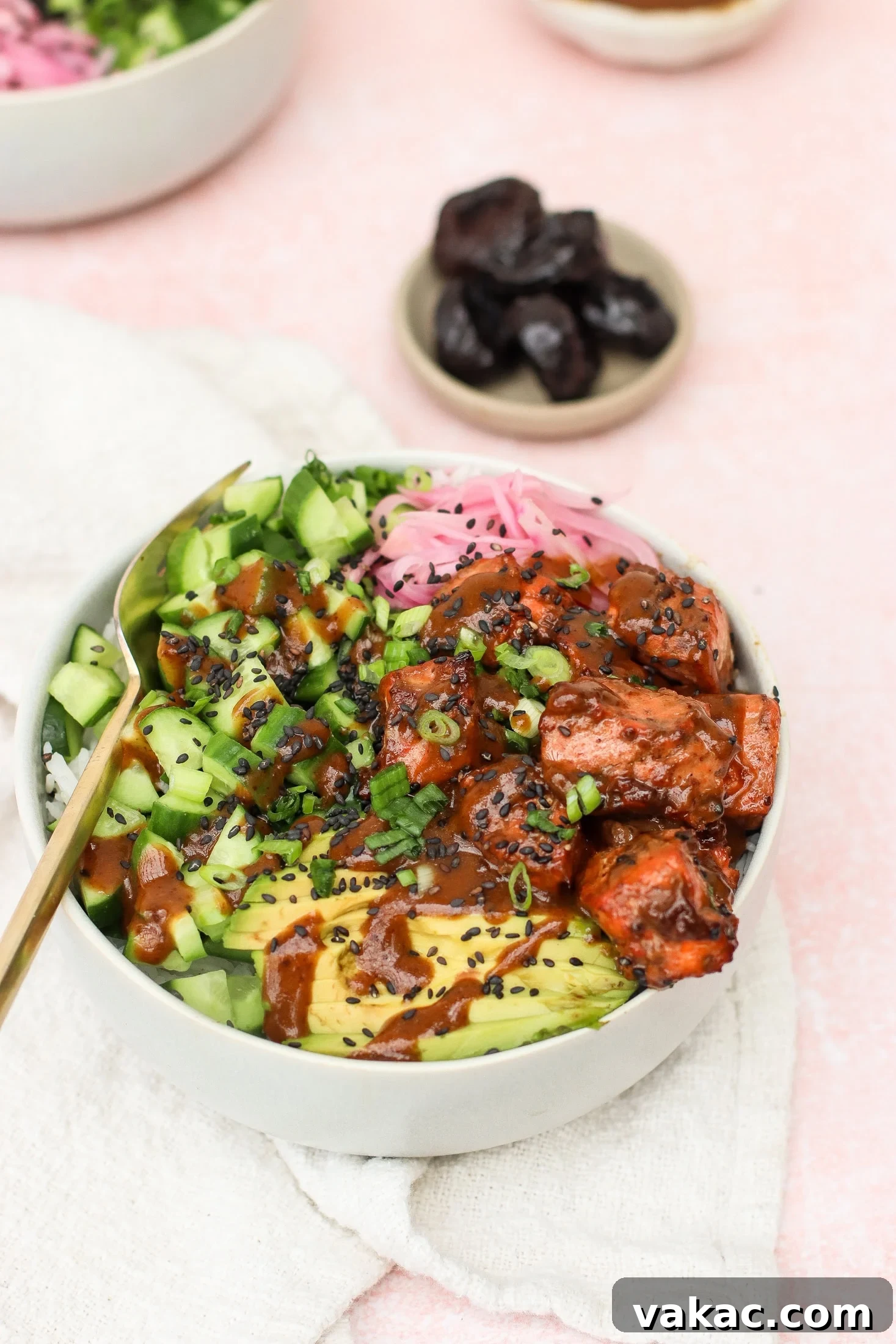 Quick Air Fryer Salmon Bowls: Sweet and Spicy Prune Delight 2 A vibrant bowl featuring perfectly cooked salmon, sliced avocado, crisp cucumber, tangy pickled onions, fresh scallions, and a sprinkle of black sesame seeds, all drizzled with a rich sauce. A small bowl of California prunes sits in the background.