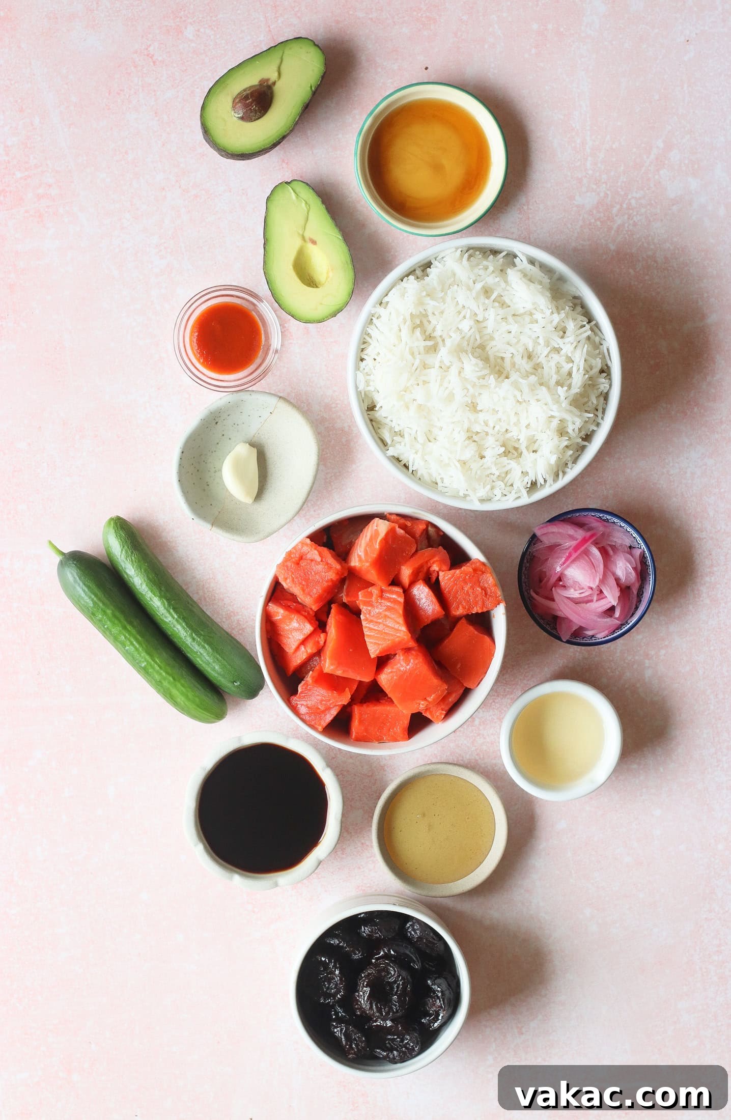 Quick Air Fryer Salmon Bowls: Sweet and Spicy Prune Delight 3 A vibrant flat lay of ingredients artfully arranged on a light pink surface, including fluffy white rice, perfectly cubed raw salmon, fresh cucumber slices, ripe avocado halves, a bottle of soy sauce, plump California prunes, a clove of garlic, a bowl of tangy pickled onions, a bottle of hot sauce, and small bowls filled with various delectable sauces, ready for preparation.