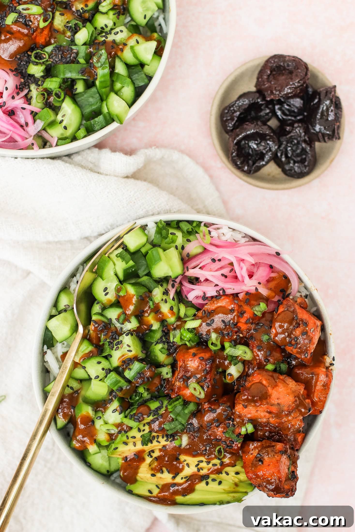 Quick Air Fryer Salmon Bowls: Sweet and Spicy Prune Delight 4 A beautifully composed bowl filled with fluffy rice, crisp diced cucumber, vibrant pickled onions, creamy avocado slices, perfectly marinated tofu (or salmon in this context), fresh green onions, and a sprinkle of black sesame seeds. A small plate of dried prunes rests nearby on a light pink surface, highlighting a key ingredient.