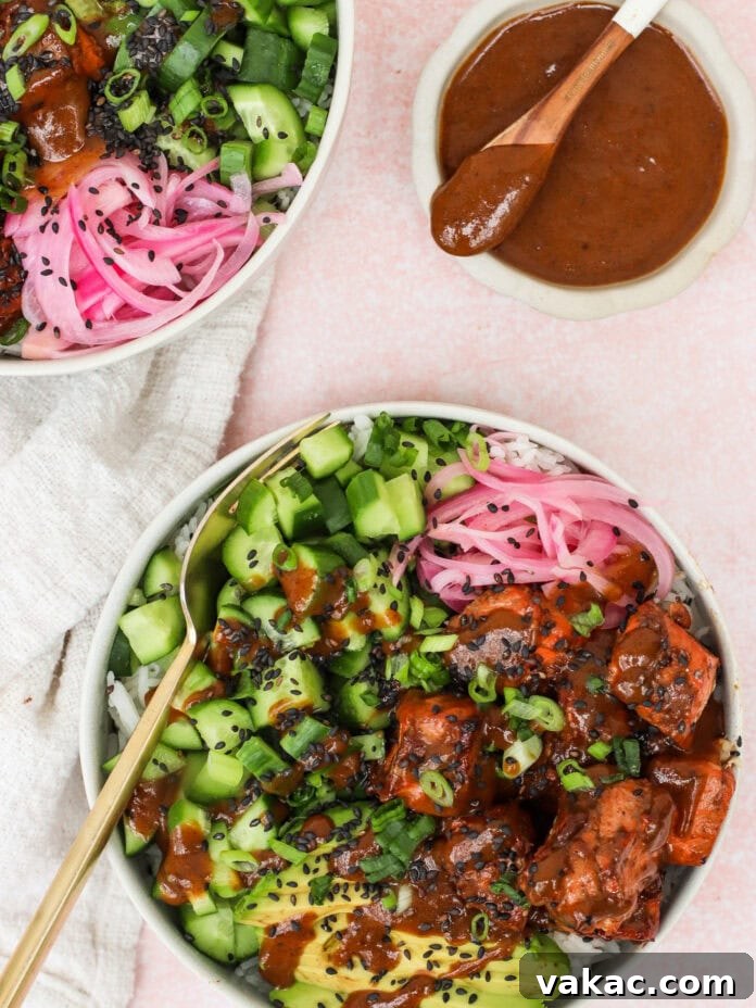 Quick Air Fryer Salmon Bowls: Sweet and Spicy Prune Delight 10 A vibrant and artfully presented rice bowl topped with perfectly glazed salmon, creamy sliced avocado, refreshing chopped cucumber, tangy pickled onions, a sprinkle of black sesame seeds, and fresh green onions. A small bowl of rich brown sauce and a spoon are neatly placed on the side, inviting you to enjoy.
