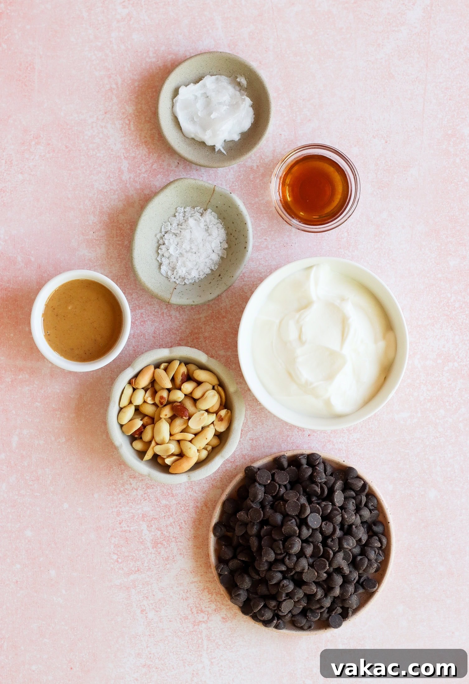 Creamy Chocolate Peanut Butter Fro Yo Treats 3 Seven small bowls on a pink surface, containing chocolate chips, peanuts, creamy yogurt, almond butter, coconut oil, flaky sea salt, and a small glass of vanilla extract.
