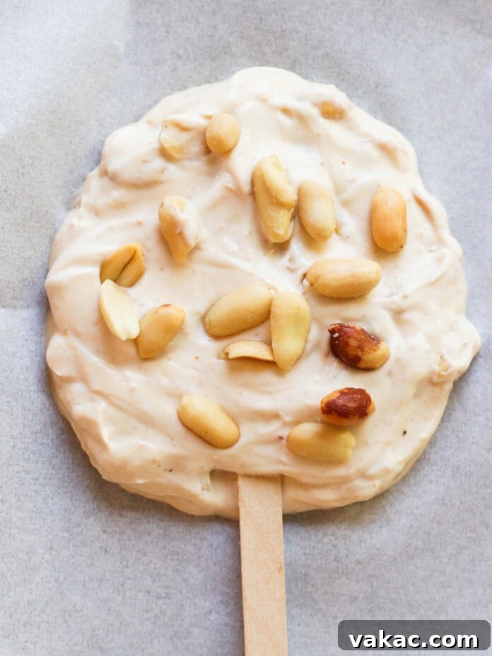 Creamy Chocolate Peanut Butter Fro Yo Treats 6 A round, creamy frozen treat on a wooden stick, topped with whole peanuts, sits on a sheet of parchment paper.
