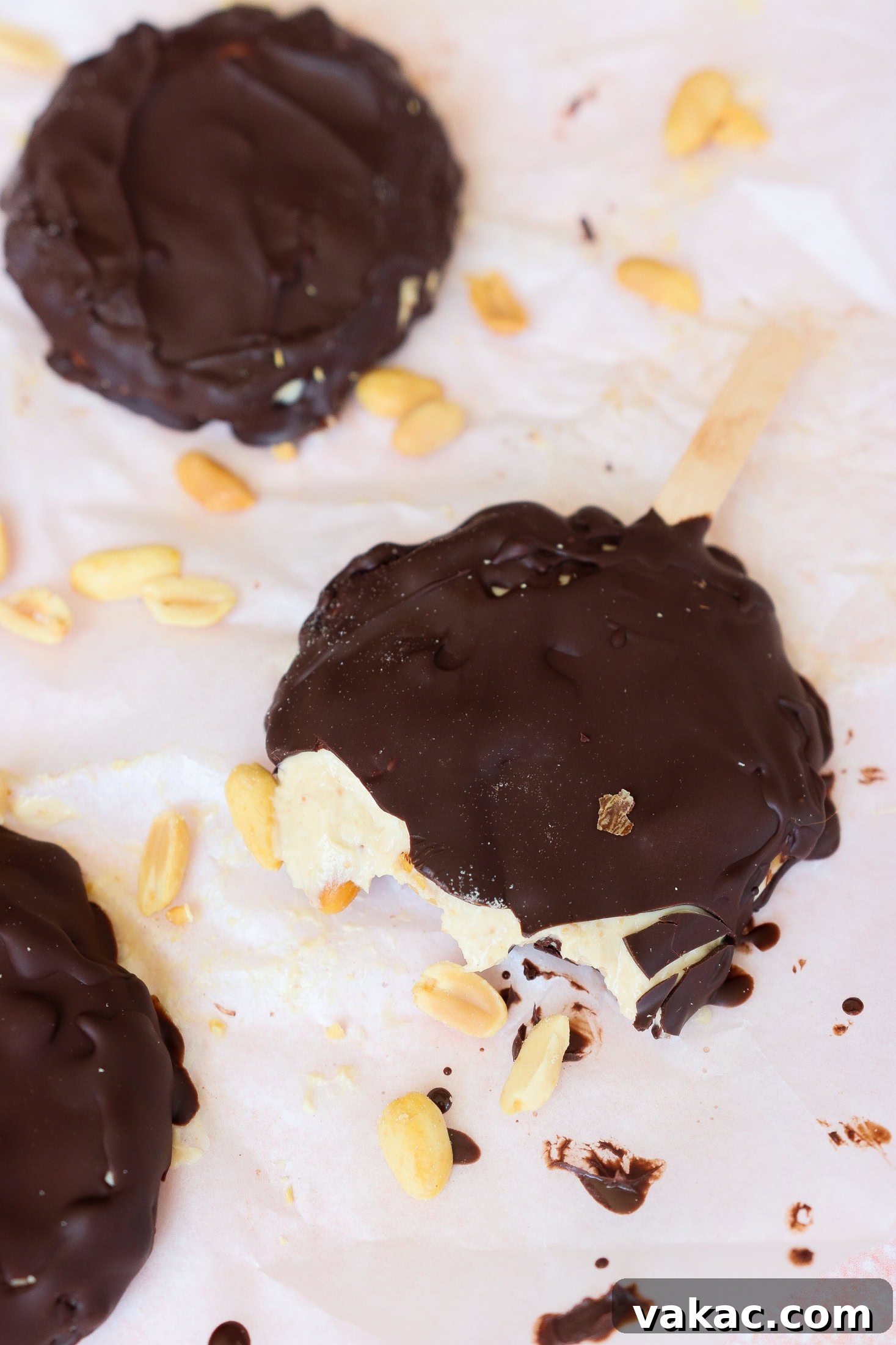Creamy Chocolate Peanut Butter Fro Yo Treats 8 Chocolate-covered ice cream pops with peanuts scattered around. One pop has a bite taken out, revealing creamy ice cream inside. The treats are on white parchment paper with melted chocolate spots.