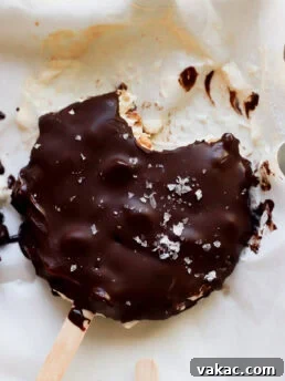 A chocolate-covered ice cream bar with a bite taken out, sprinkled with flaky sea salt, rests on parchment paper with some melted chocolate and ice cream visible around it.