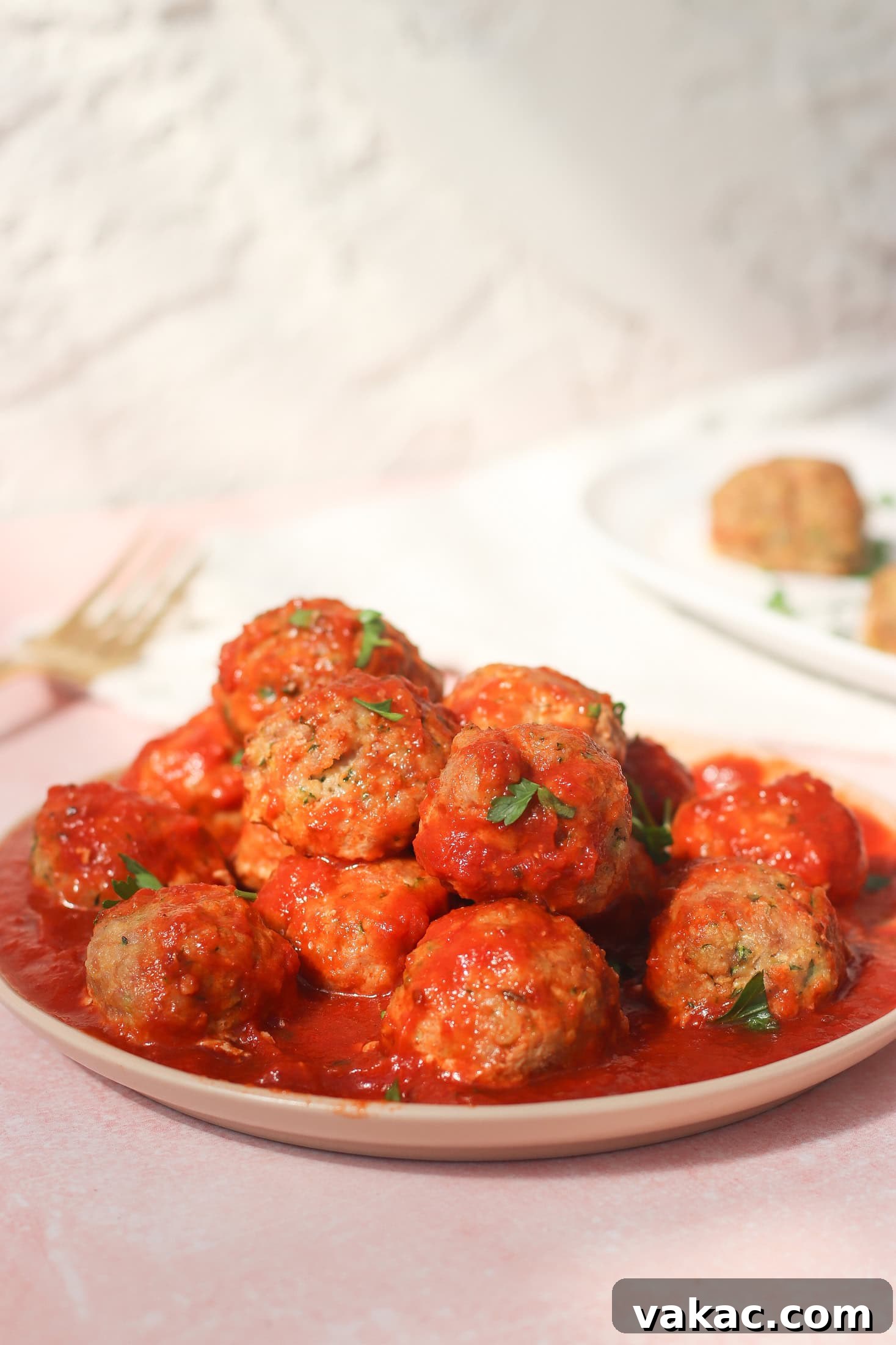 Quick Sheet Pan Turkey Zucchini Meatballs Tender and Juicy 2 A plate of baked turkey and zucchini meatballs covered in a rich tomato sauce, garnished with fresh chopped parsley, sits on a light pink surface with a fork and another plate of food blurred in the background. The scene suggests a cozy, delicious meal ready to be enjoyed.