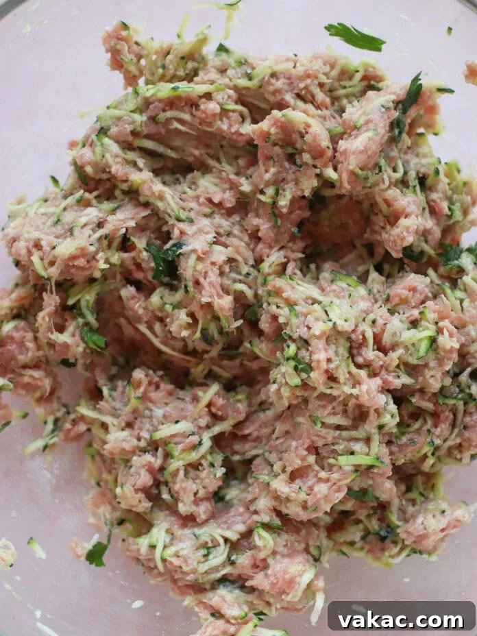 Quick Sheet Pan Turkey Zucchini Meatballs Tender and Juicy 4 A close-up view of raw ground turkey mixed with shredded zucchini, fresh herbs, and other seasonings in a clear glass mixing bowl, ready for the next step of the recipe.