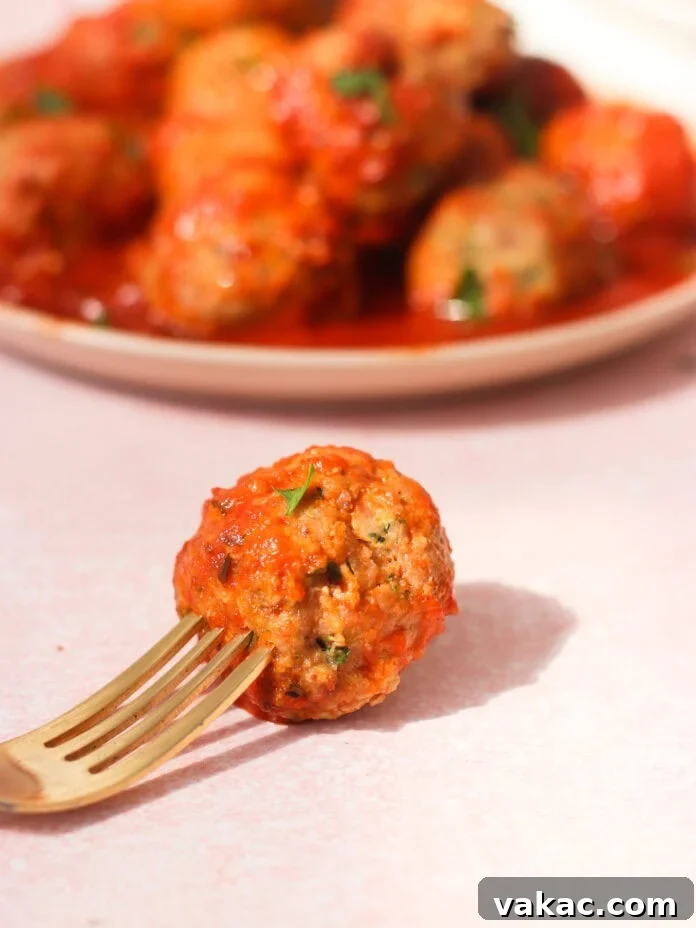 Quick Sheet Pan Turkey Zucchini Meatballs Tender and Juicy 7 A golden fork holds a single, perfectly cooked turkey zucchini meatball, generously coated in a rich tomato sauce. In the softly blurred background, a plate of more saucy meatballs awaits, all resting on a light pink surface, emphasizing the deliciousness of the meal.