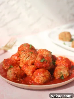 Quick Sheet Pan Turkey Zucchini Meatballs Tender and Juicy 8 A plate of meatballs covered in tomato sauce, garnished with chopped parsley, sits on a light pink surface with a fork and another plate of food blurred in the background.