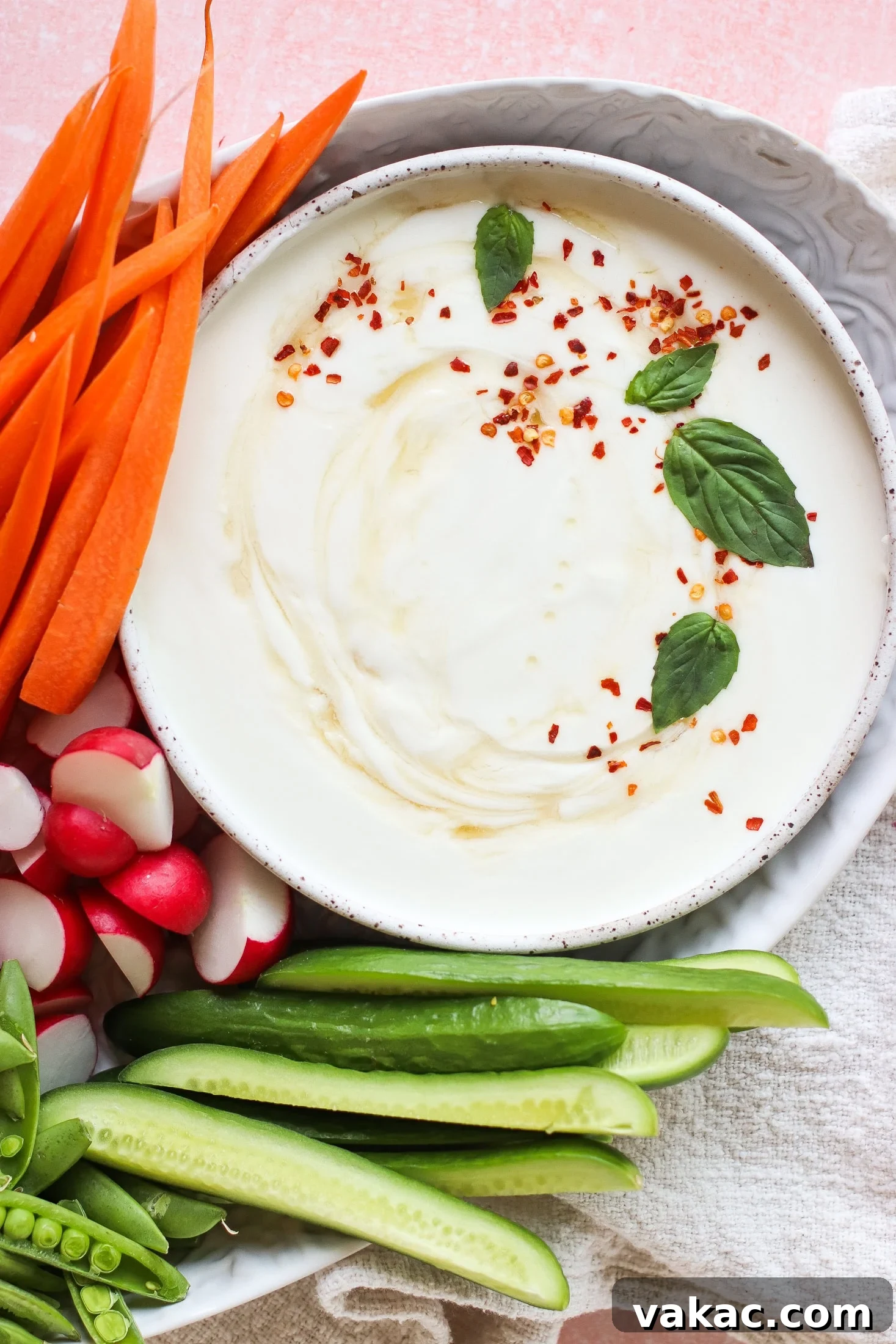 A vibrant bowl of creamy whipped cottage cheese dip, elegantly garnished with red pepper flakes and fresh basil leaves. It's artfully arranged amidst a selection of crisp, colorful vegetables including bright carrot sticks, delicate radish halves, cool cucumber slices, and sweet snap peas, inviting a wholesome and flavorful experience.