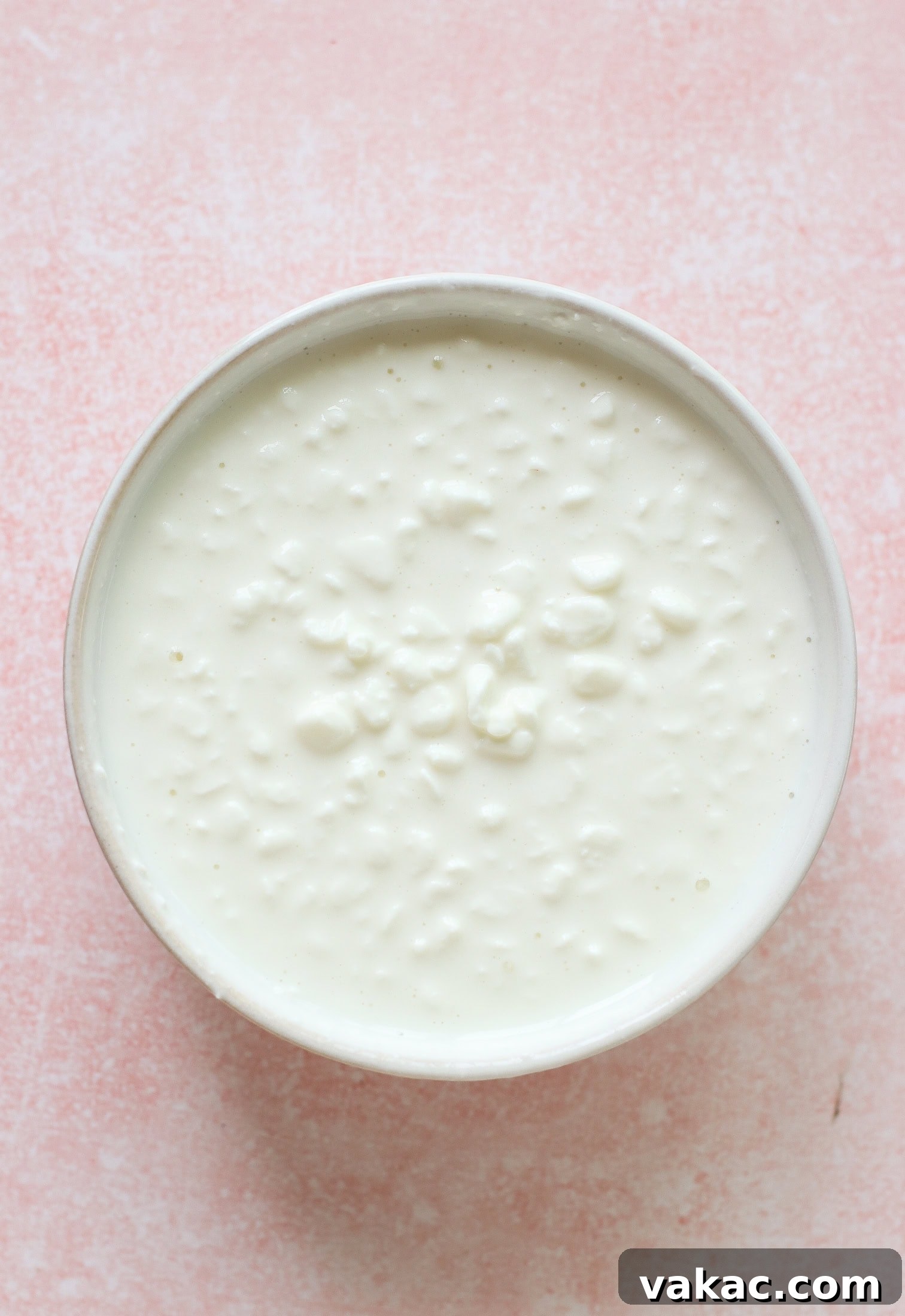 A pristine, overhead view of a bowl filled with small-curd cottage cheese, resting on a textured light pink surface. The creamy, white curds are clearly visible, indicating the fresh and simple nature of the ingredient.