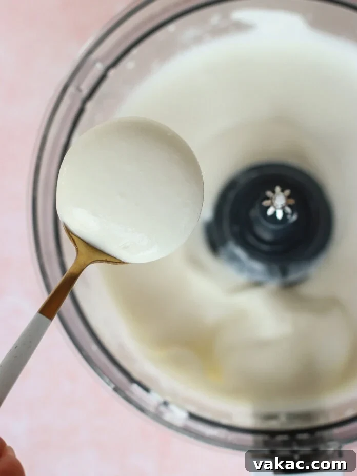 A close-up shot of a spoon expertly scooping a generous portion of thick, creamy white yogurt from an open food processor bowl, which is also filled with the same velvety mixture. This image captures the rich texture and readiness of the blended product.