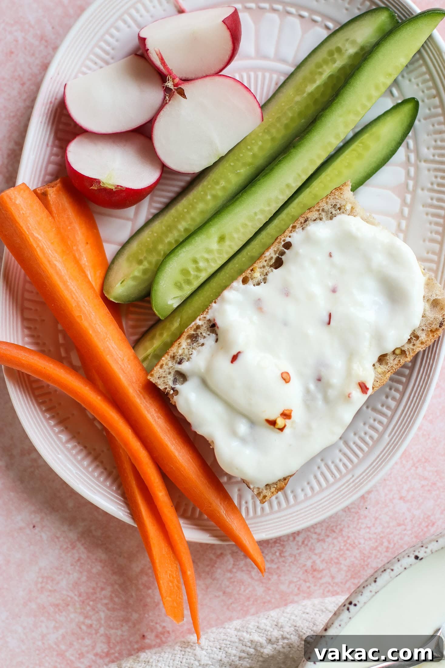 A thoughtfully composed plate featuring an array of fresh, vibrant components. It holds thinly sliced radishes, crisp cucumber spears, bright orange carrot sticks, and a piece of crispbread generously topped with a creamy white spread, artfully finished with a sprinkle of chili flakes. This tableau emphasizes healthy snacking and balanced textures.