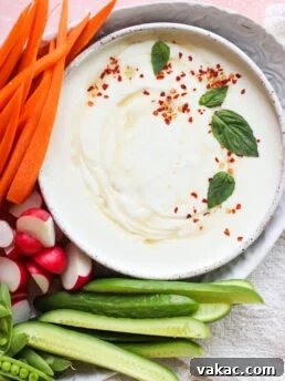 A bowl of creamy cottage cheese dip garnished with red pepper flakes and basil leaves is surrounded by fresh vegetables: carrot sticks, radish halves, cucumber slices, and snap peas.