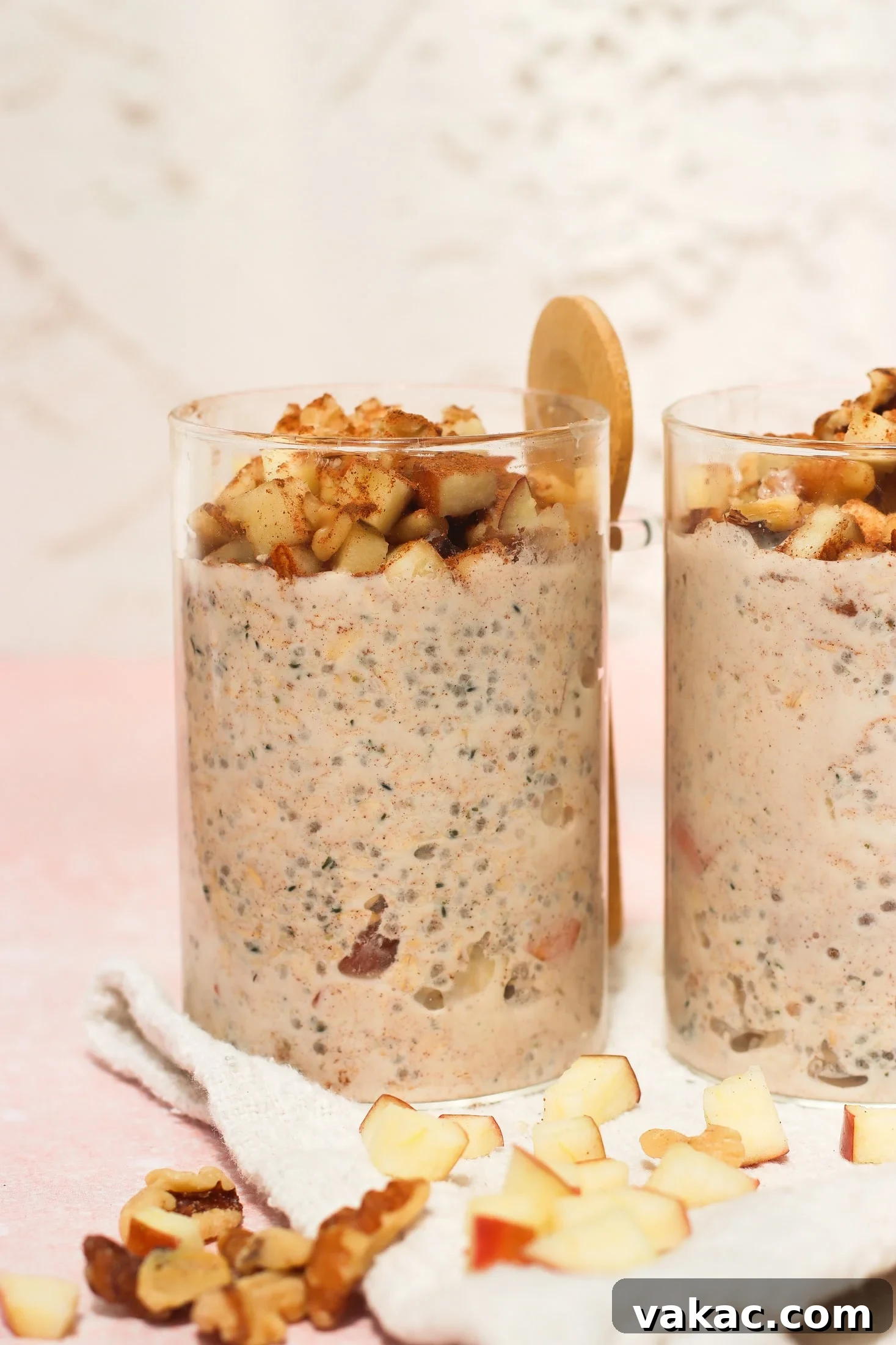 Protein-Packed Apple Cinnamon Overnight Oats 2 Two clear glasses filled with a creamy oatmeal mixture topped with chopped apples and walnuts. A wooden spoon rests behind one glass. Diced apples and walnuts are scattered on a white cloth beneath the glasses.