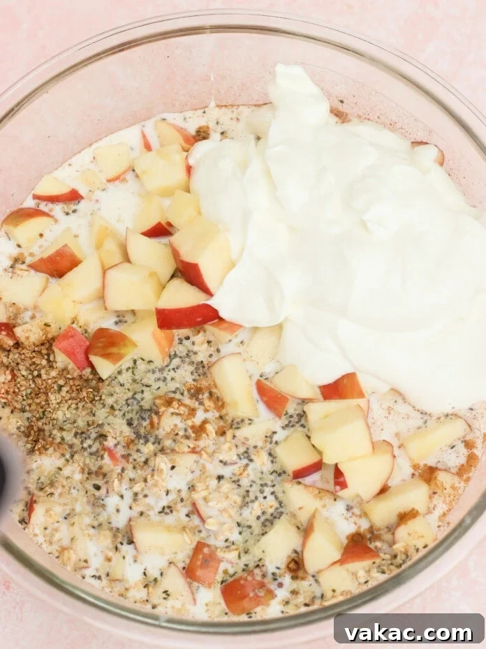 Protein-Packed Apple Cinnamon Overnight Oats 4 A glass bowl containing chopped apples, yogurt, seeds, spices, and milk on a pink surface, ready to be mixed for a recipe.