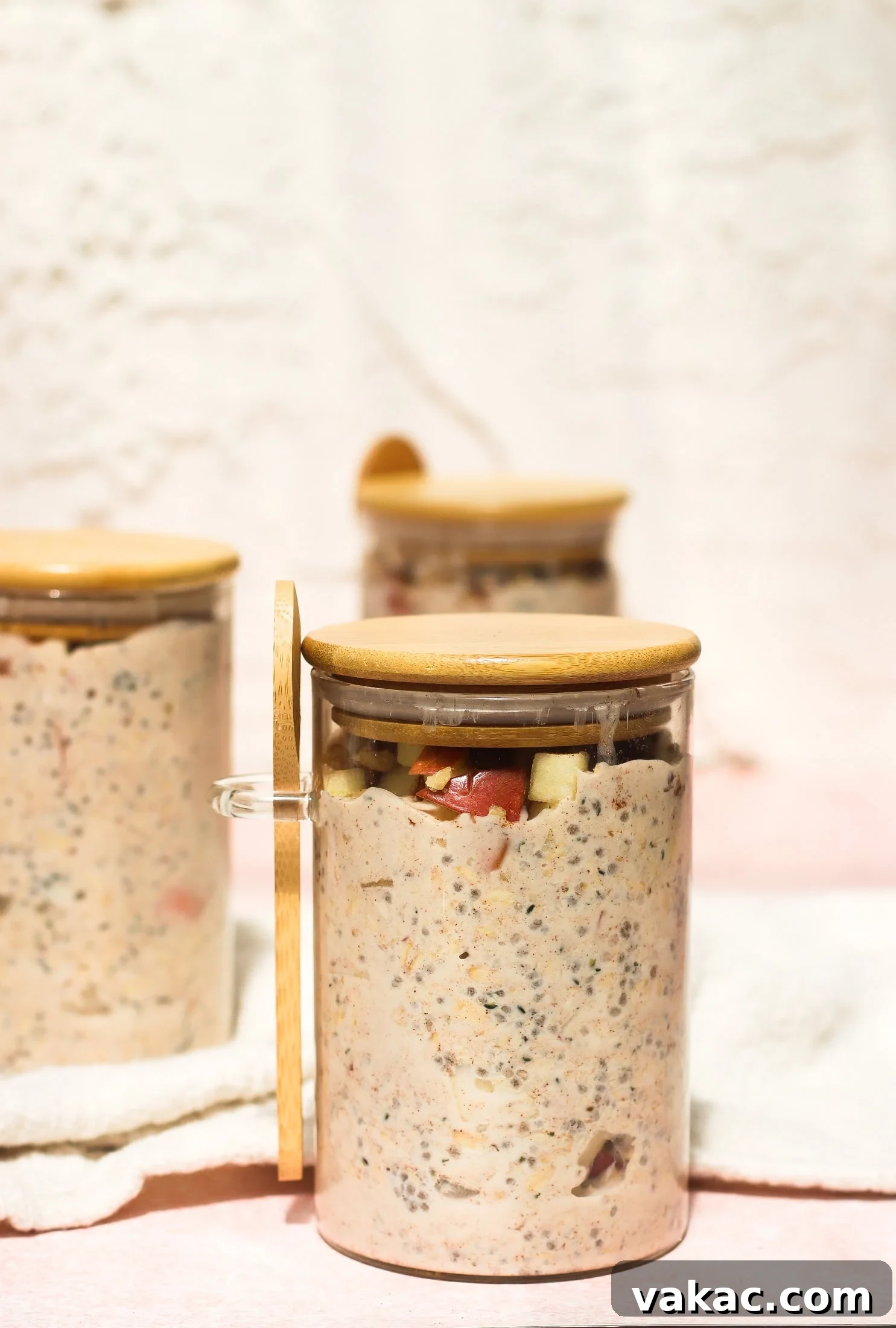Protein-Packed Apple Cinnamon Overnight Oats 6 Three glass jars with wooden lids hold a creamy mixture containing visible fruit and chia seeds. The jar in the foreground is open, showing the contents more clearly. The background is softly lit and out of focus.