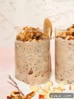 Two clear glasses filled with a creamy oatmeal mixture topped with chopped apples and walnuts. A wooden spoon rests behind one glass. Diced apples and walnuts are scattered on a white cloth beneath the glasses.