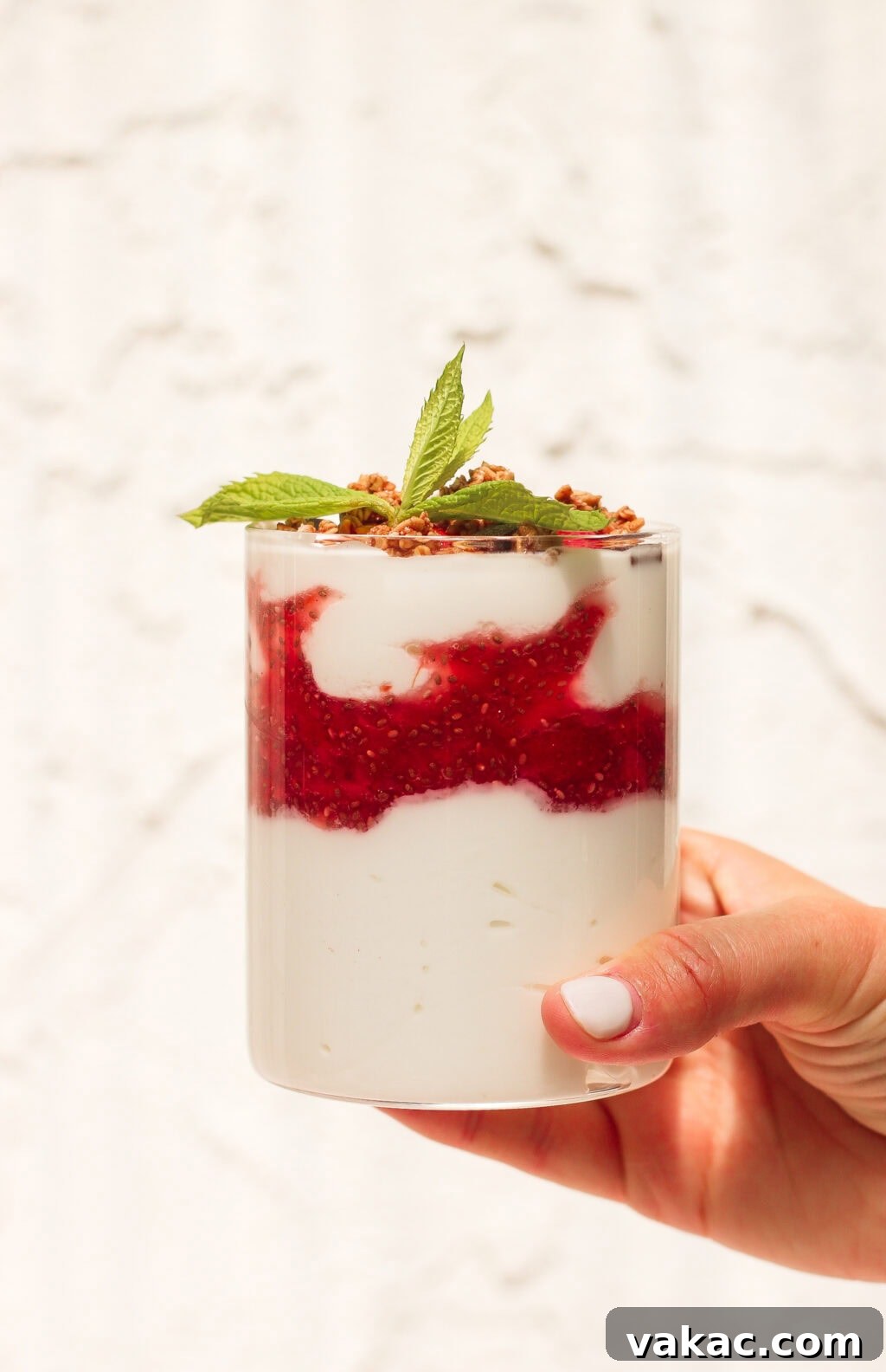Instant Strawberry Greek Yogurt Stack 2 A hand holds a glass filled with layered yogurt, red berry compote, granola, and topped with a sprig of mint against a light, textured background, highlighting the vibrant colors and fresh appeal of the parfait.
