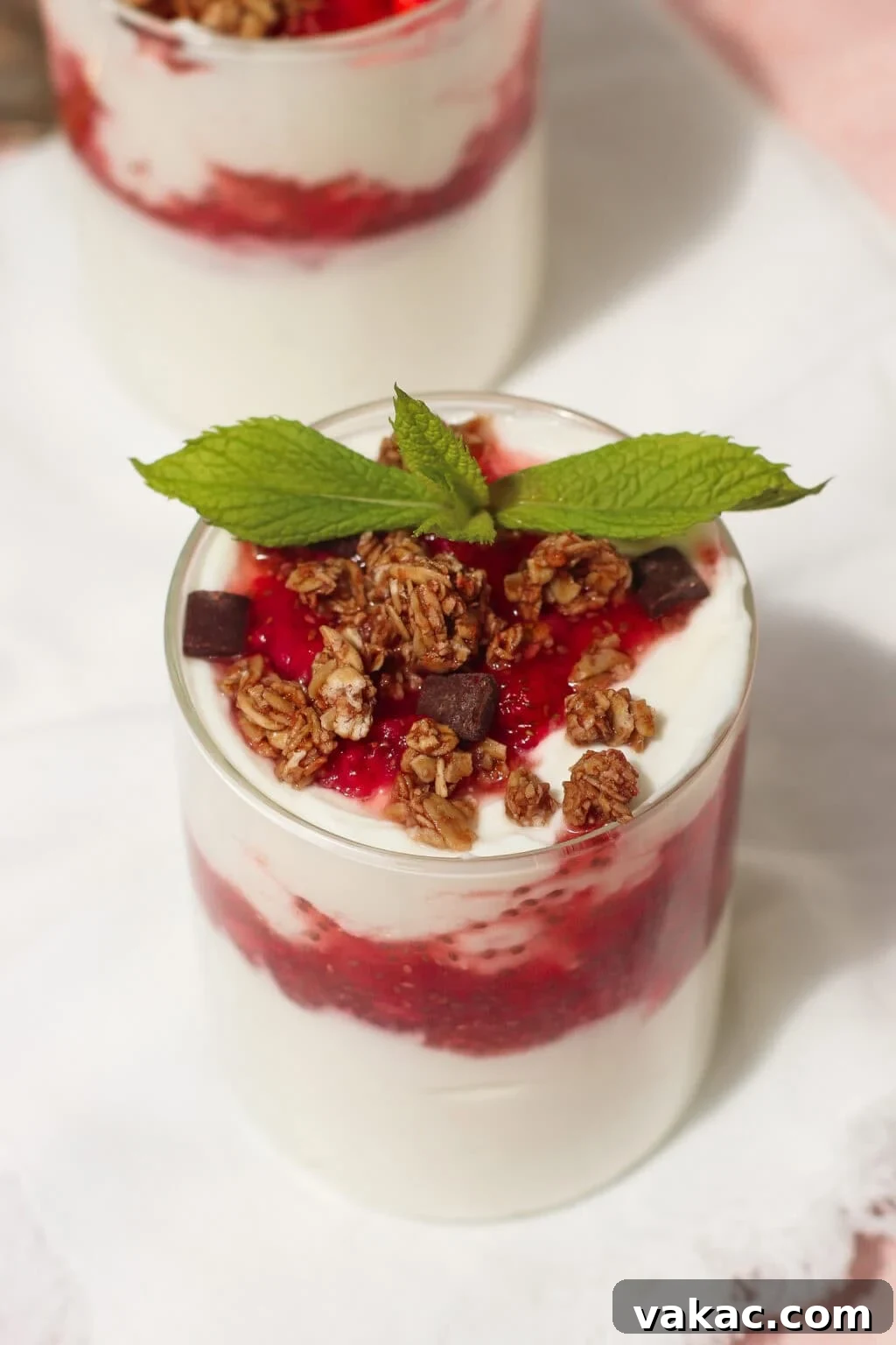 Instant Strawberry Greek Yogurt Stack 3 A beautifully presented glass of yogurt parfait, artfully layered with a rich red berry sauce, topped with crunchy granola, tempting chocolate chunks, and a fresh sprig of mint, all elegantly placed on a pristine white napkin, inviting a delightful breakfast experience.