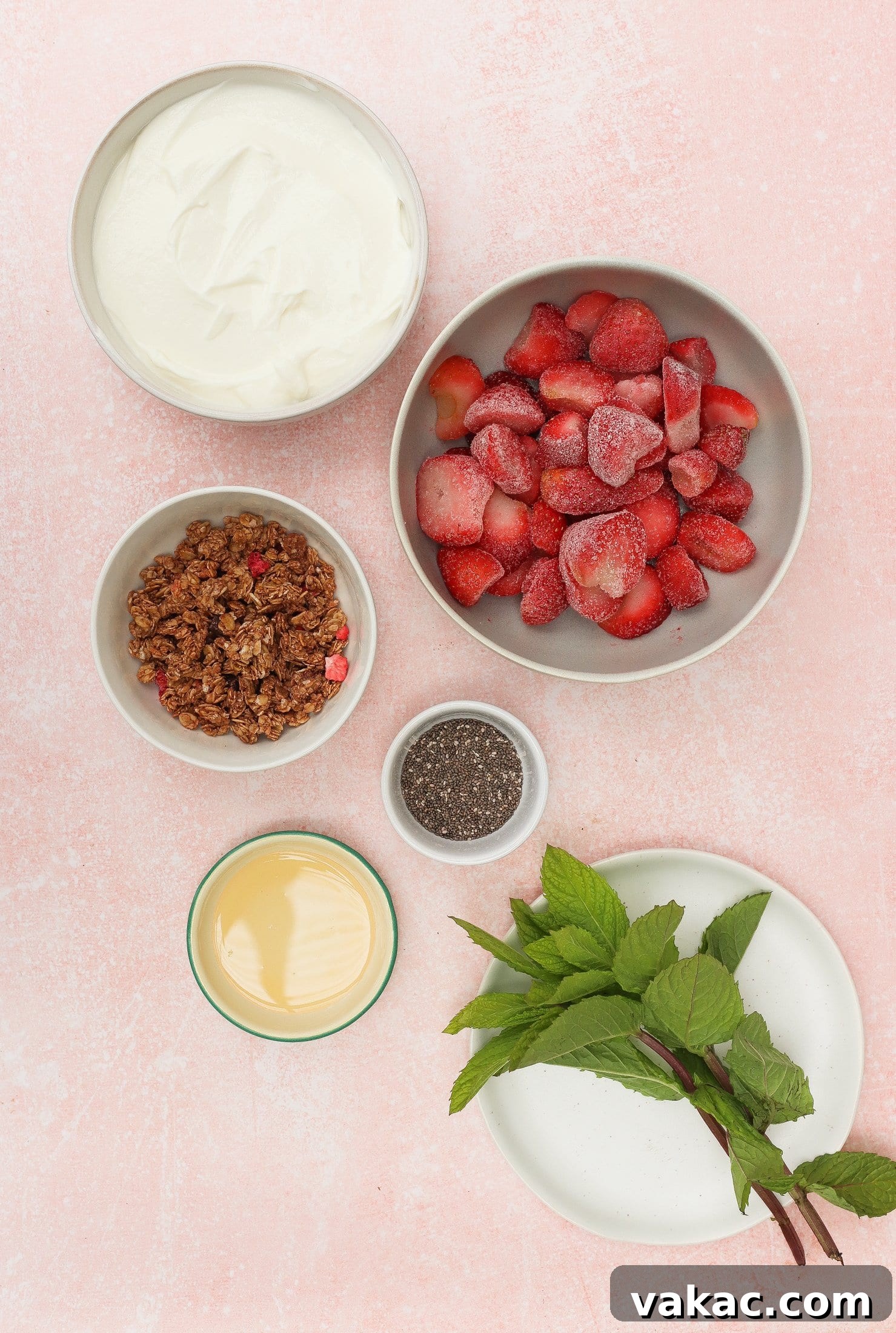 Instant Strawberry Greek Yogurt Stack 4 A vibrant flat lay shows five distinct bowls arranged on a soft pink surface, each containing a key ingredient for the parfait: fluffy whipped cream, juicy frozen strawberries, crunchy granola, tiny chia seeds, and glistening sweetened condensed milk, with sprigs of fresh mint on a white plate nearby, ready to garnish.