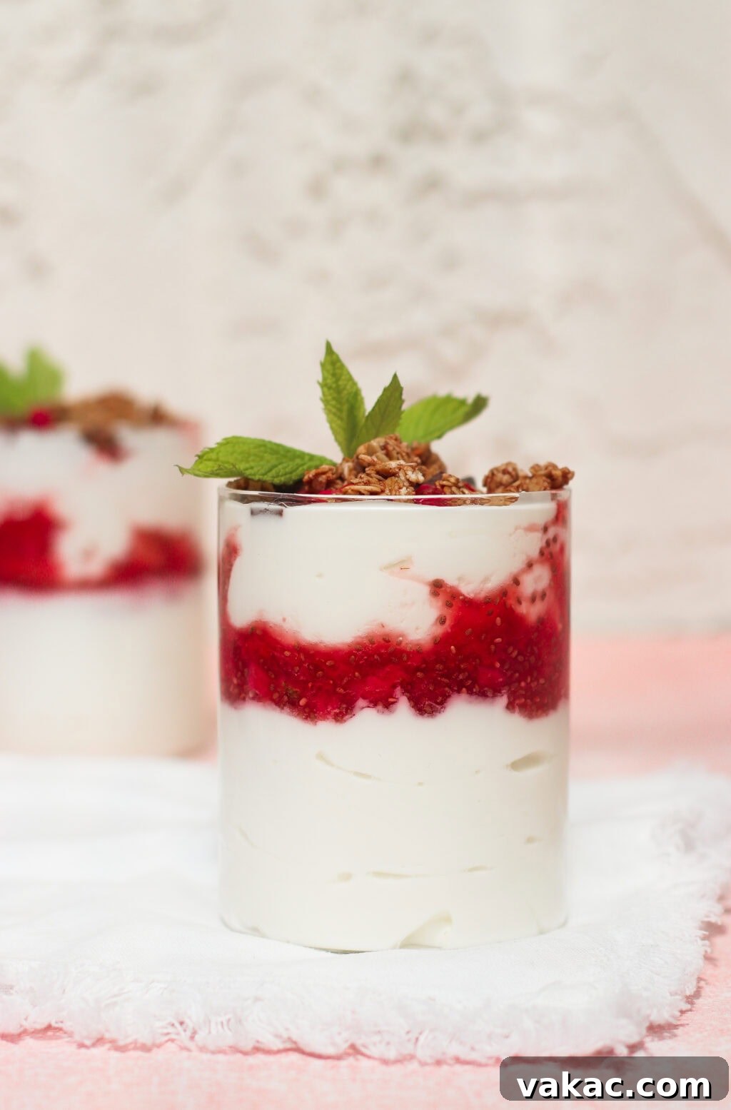 Instant Strawberry Greek Yogurt Stack 7 A pristine glass filled with layers of creamy yogurt and a vibrant red berry sauce, generously topped with crunchy granola and a fresh sprig of mint, sits on a crisp white napkin, with another identical dessert blurred elegantly in the background, suggesting a delightful pair of treats.