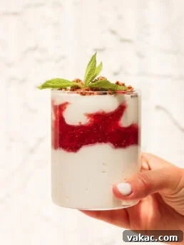 A hand holds a glass filled with layered yogurt, red berry compote, granola, and topped with a sprig of mint against a light, textured background, ready to be enjoyed.