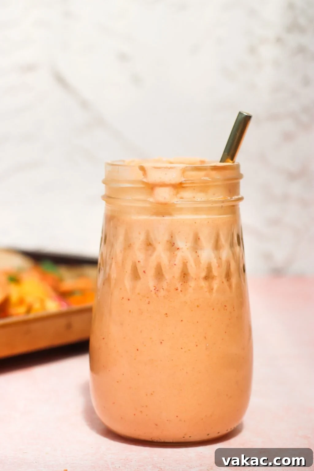 Velvety Greek Yogurt Chipotle Sauce Ready in Minutes 2 A mason jar filled with a creamy, light orange smoothie and topped with froth, with a metal straw inside. The jar sits on a light pink surface, and the background is softly blurred.