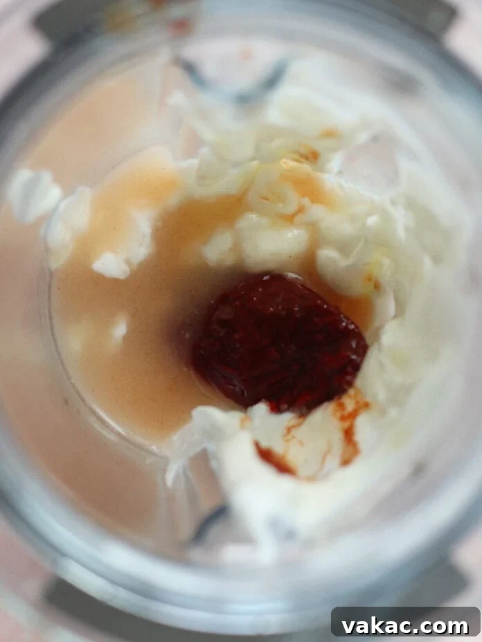Velvety Greek Yogurt Chipotle Sauce Ready in Minutes 3 Overhead view of a blender cup containing pale liquid, pieces of chopped white onion, and a red chili pepper or paste in the center, ready to be blended.