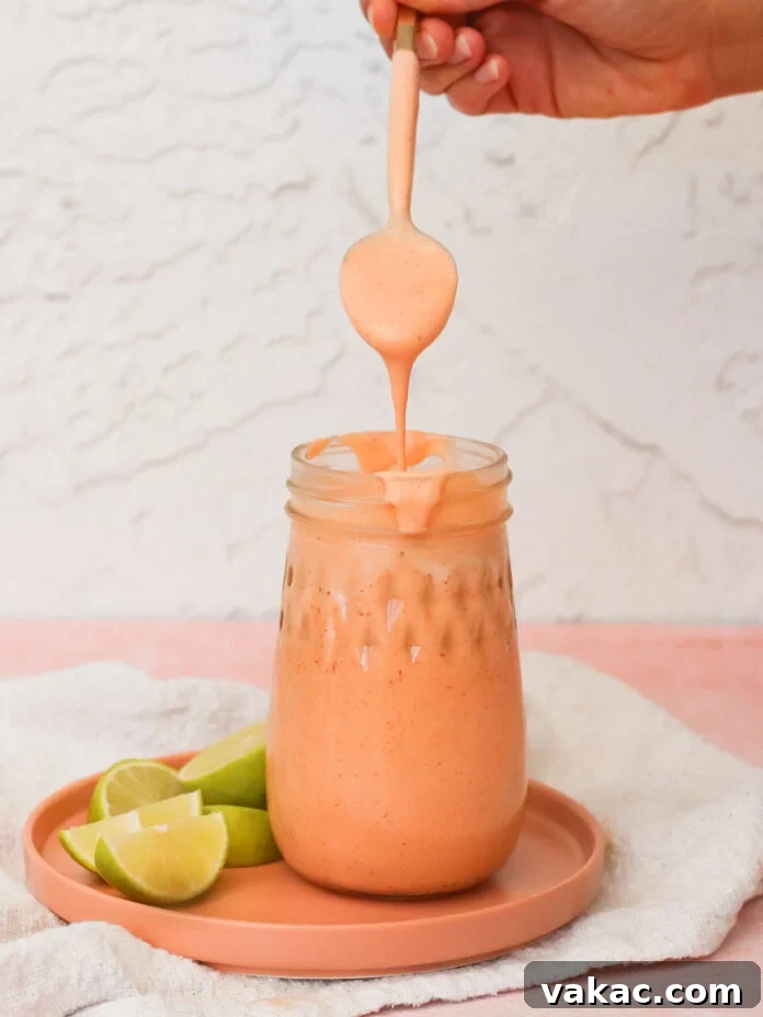 Velvety Greek Yogurt Chipotle Sauce Ready in Minutes 4 A hand holds a spoon drizzling thick, creamy, orange sauce into a mason jar on a peach-colored plate with lime wedges beside it, against a light textured background.