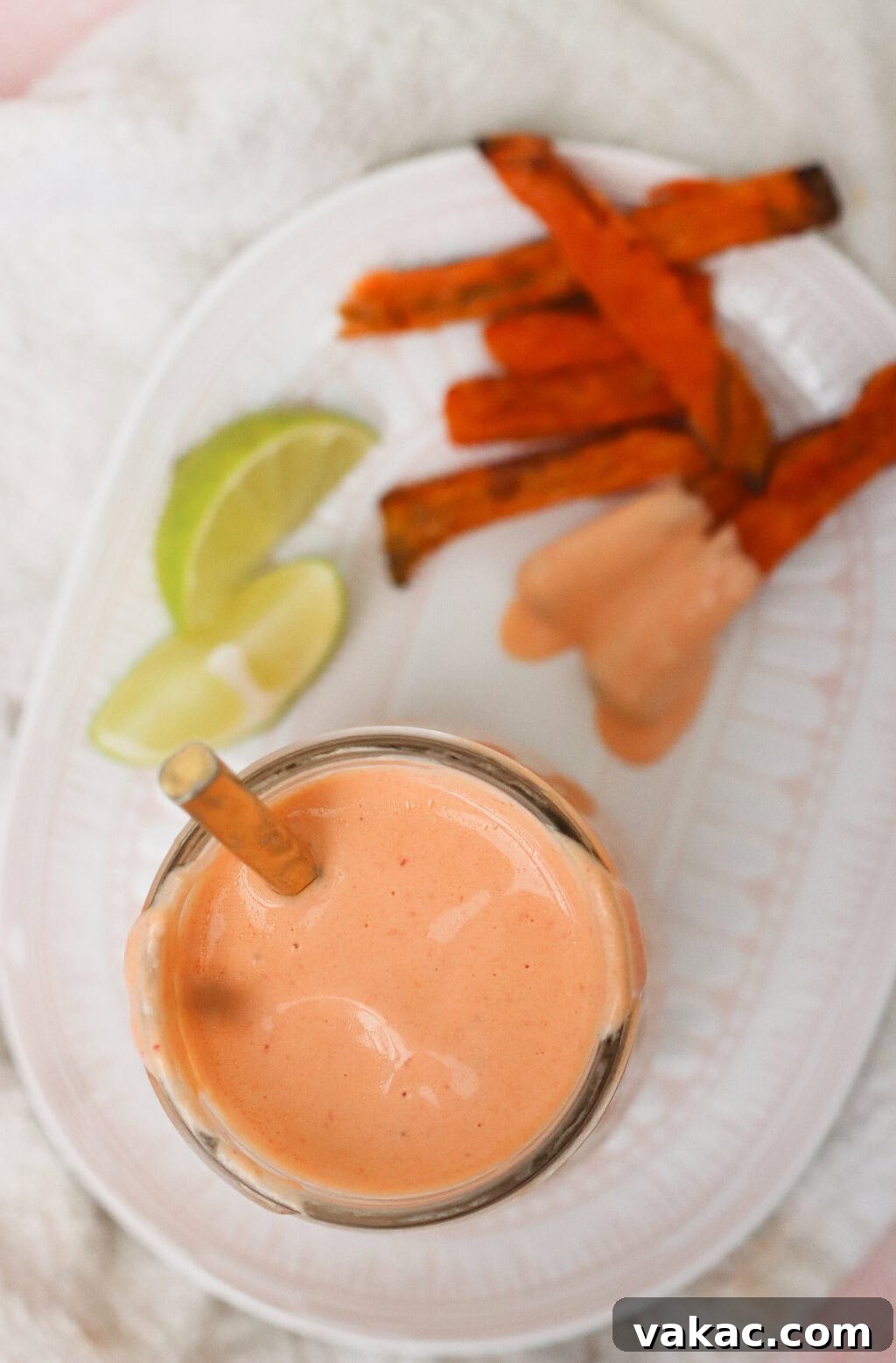 Velvety Greek Yogurt Chipotle Sauce Ready in Minutes 5 A glass of creamy pink dipping sauce with a spoon, next to sweet potato fries, two lime wedges, and a dollop of sauce on a white plate.