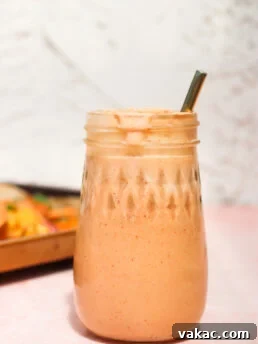 A mason jar filled with a creamy, light orange smoothie and topped with froth, with a metal straw inside. The jar sits on a light pink surface, and the background is softly blurred.