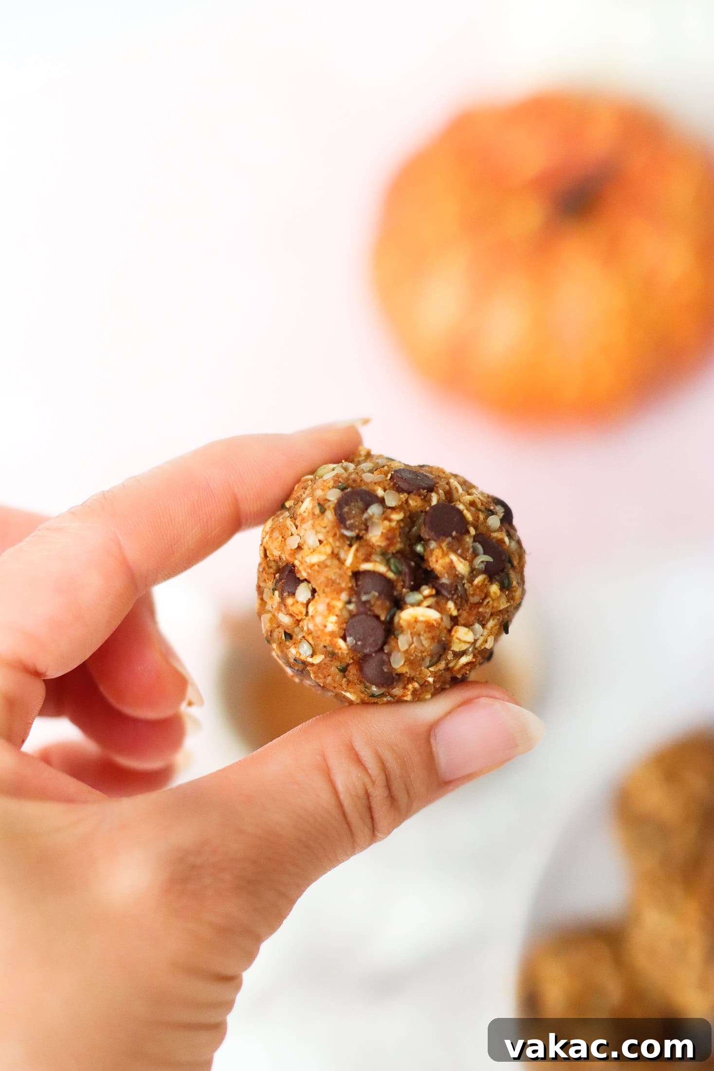 Speedy Pumpkin Spice Almond Flour Protein Bites 6 A hand holding a round, oat and chocolate chip energy bite, with a blurred pumpkin and more energy bites in the background.
