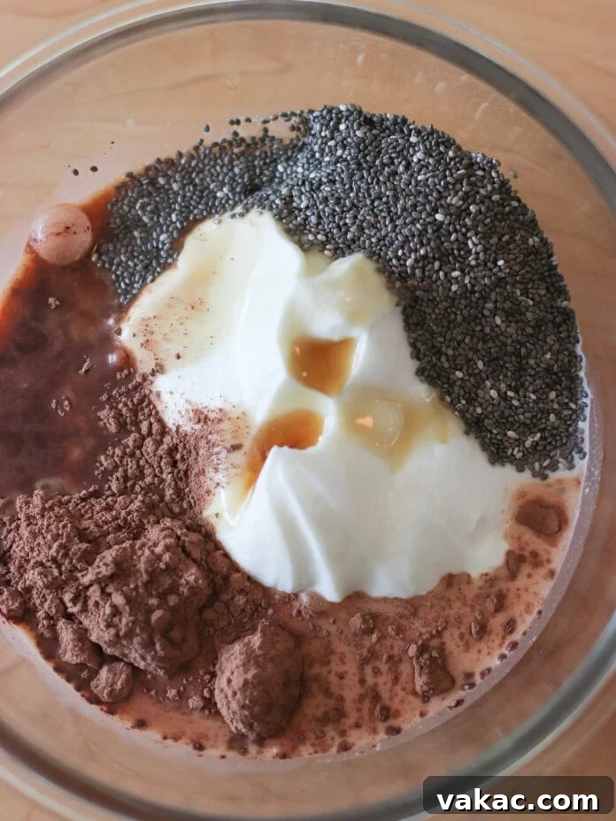 Velvet Chocolate Chia Protein Pudding with Greek Yogurt 4 A glass bowl containing yogurt, cocoa powder, chia seeds, vanilla extract, and milk, all unmixed, on a wooden surface.