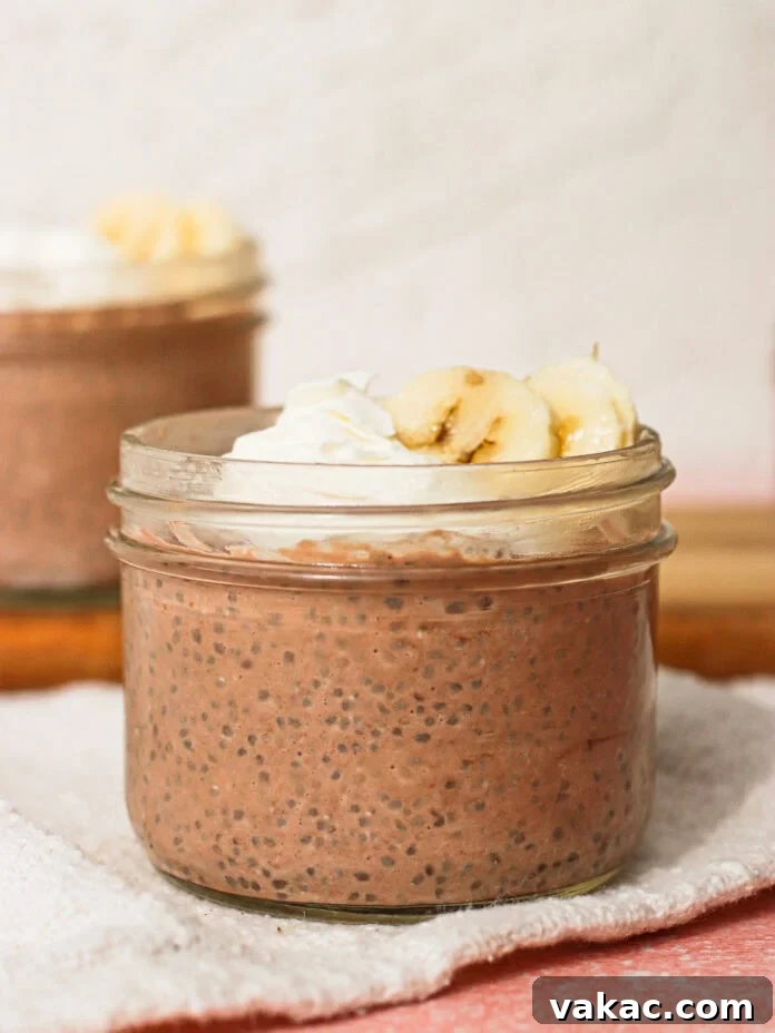Velvet Chocolate Chia Protein Pudding with Greek Yogurt 7 A small glass jar filled with chocolate chia pudding, topped with whipped cream and banana slices, sits on a white cloth. Another similar jar is blurred in the background.