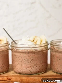 Three glass jars filled with chocolate chia pudding, topped with banana slices and whipped cream, are displayed on a wooden board. One jar has a spoon inside. The background is light and neutral.