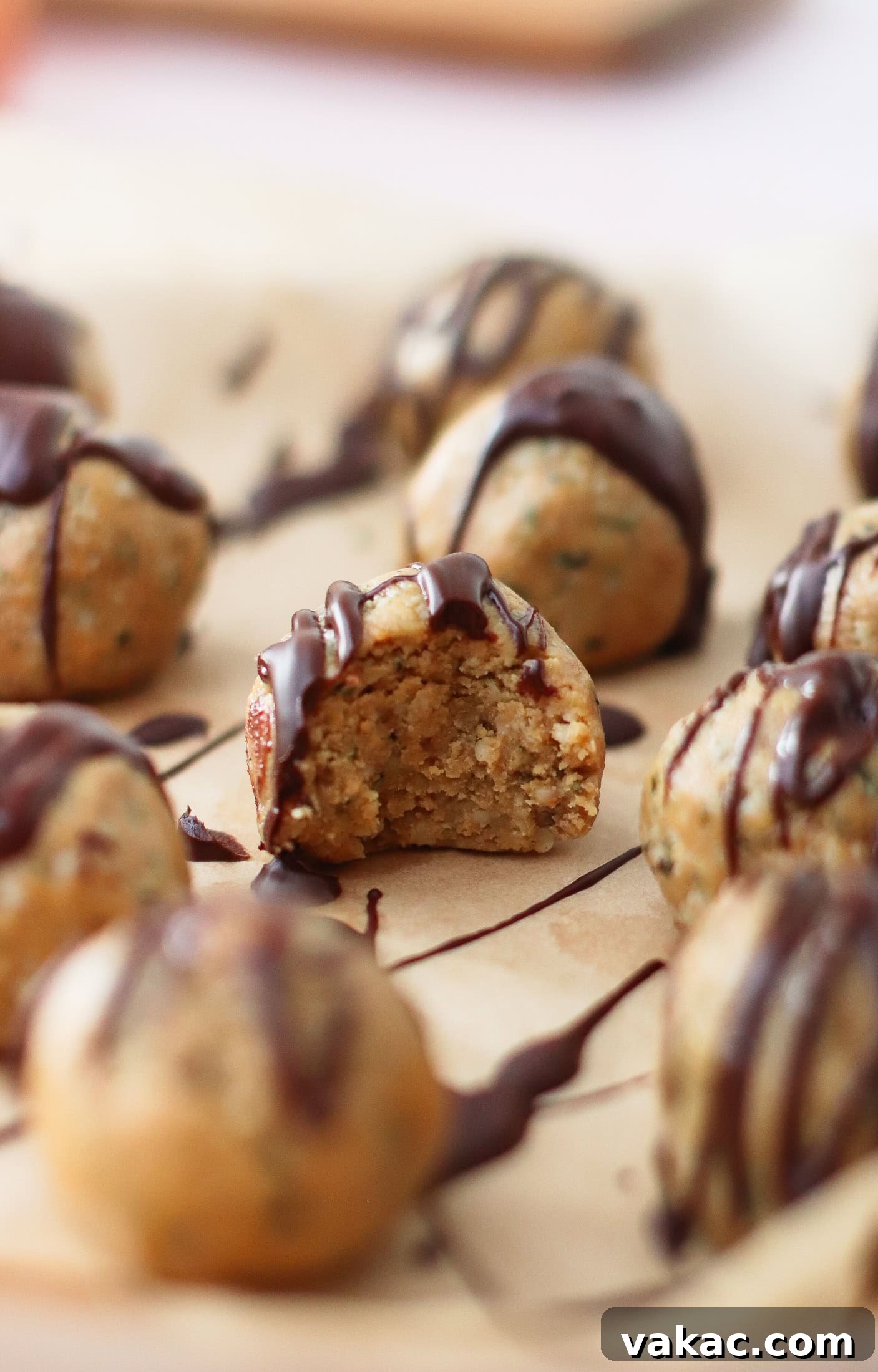 No-Bake Peanut Butter Oat Flour Energy Bites 8 Close-up of round, cookie dough bites drizzled with chocolate on parchment paper. The center bite is cut in half, showing a soft, textured interior.