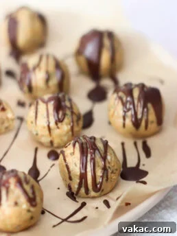 Round, bite-sized cookie balls on parchment paper are drizzled with dark chocolate. The treats appear homemade, with a nutty, textured surface and a glossy chocolate topping. Some are in focus, while others are blurred in the background.