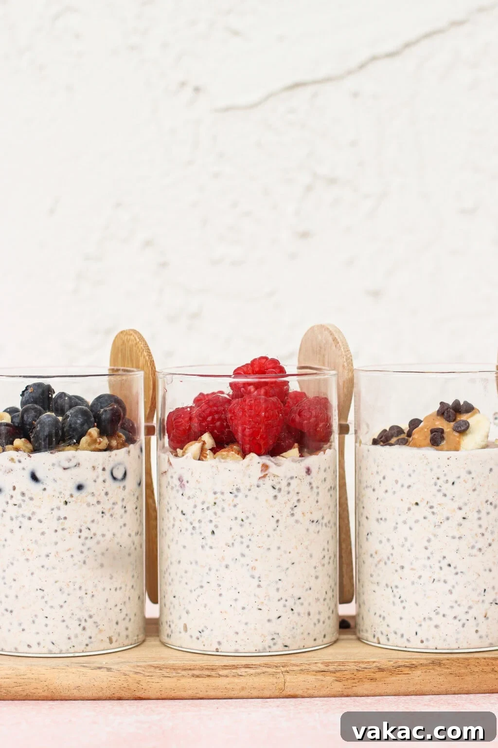 Three clear glasses filled with overnight oats are topped with blueberries, raspberries, and banana slices with chocolate chips. Wooden spoons are placed behind each glass, and the background is a light textured wall.