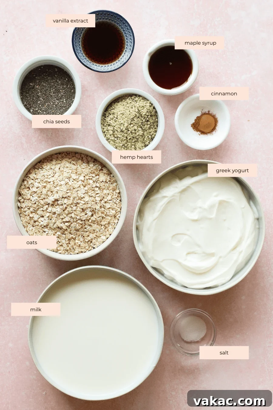 Various ingredients for a healthy breakfast are arranged on a pink surface, including labeled bowls of oats, milk, Greek yogurt, chia seeds, hemp hearts, vanilla extract, maple syrup, cinnamon, and salt.
