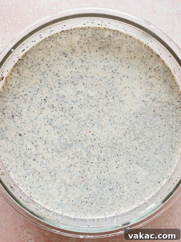 A glass bowl filled with a creamy mixture speckled with small black seeds, likely poppy seeds, sits on a light pink textured surface.