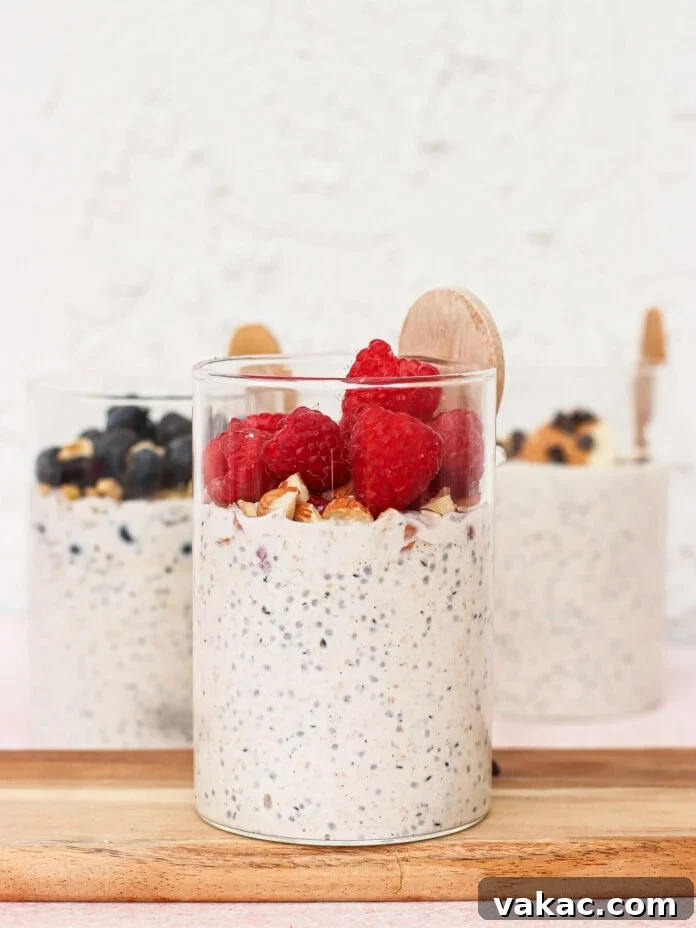 Three glass cups of overnight oats topped with fresh berries and granola sit on a wooden board. The front cup has raspberries, while the back cups have blueberries and wooden spoons in each.