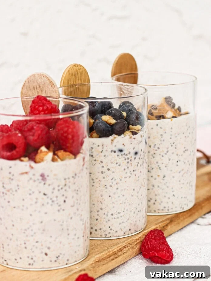 Three glasses of chia seed pudding topped with fresh raspberries, blueberries, and chopped nuts, each with a wooden spoon, displayed on a wooden board with scattered raspberries nearby.