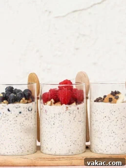 Three clear glasses filled with overnight oats are topped with blueberries, raspberries, and banana slices with chocolate chips. Wooden spoons are placed behind each glass, and the background is a light textured wall.