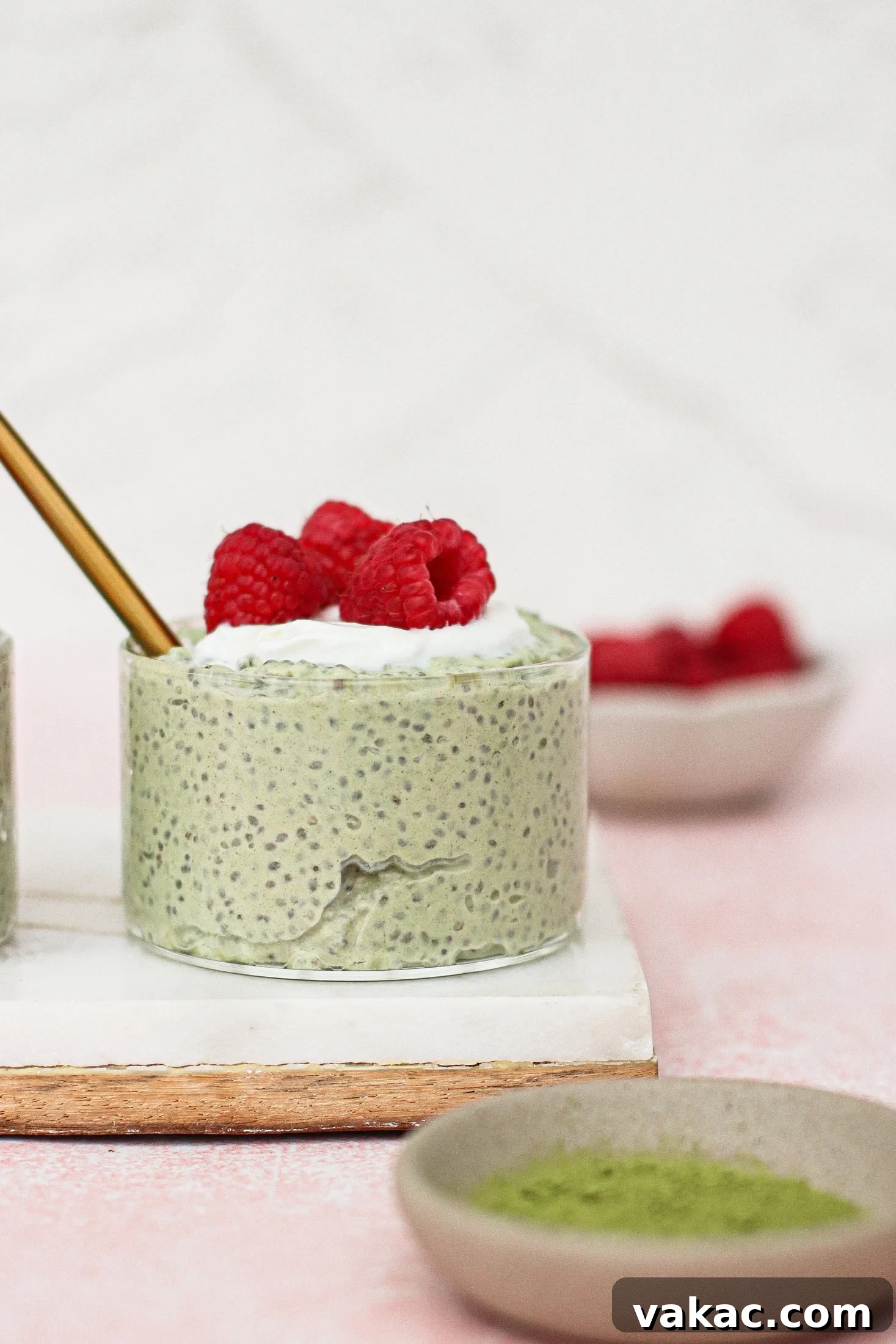 Quick High Protein Matcha Chia Pudding with Yogurt 2 A small round dish of matcha chia pudding topped with whipped cream and fresh raspberries sits on a white board. A gold spoon is in the pudding, and bowls of raspberries and green powder are nearby.