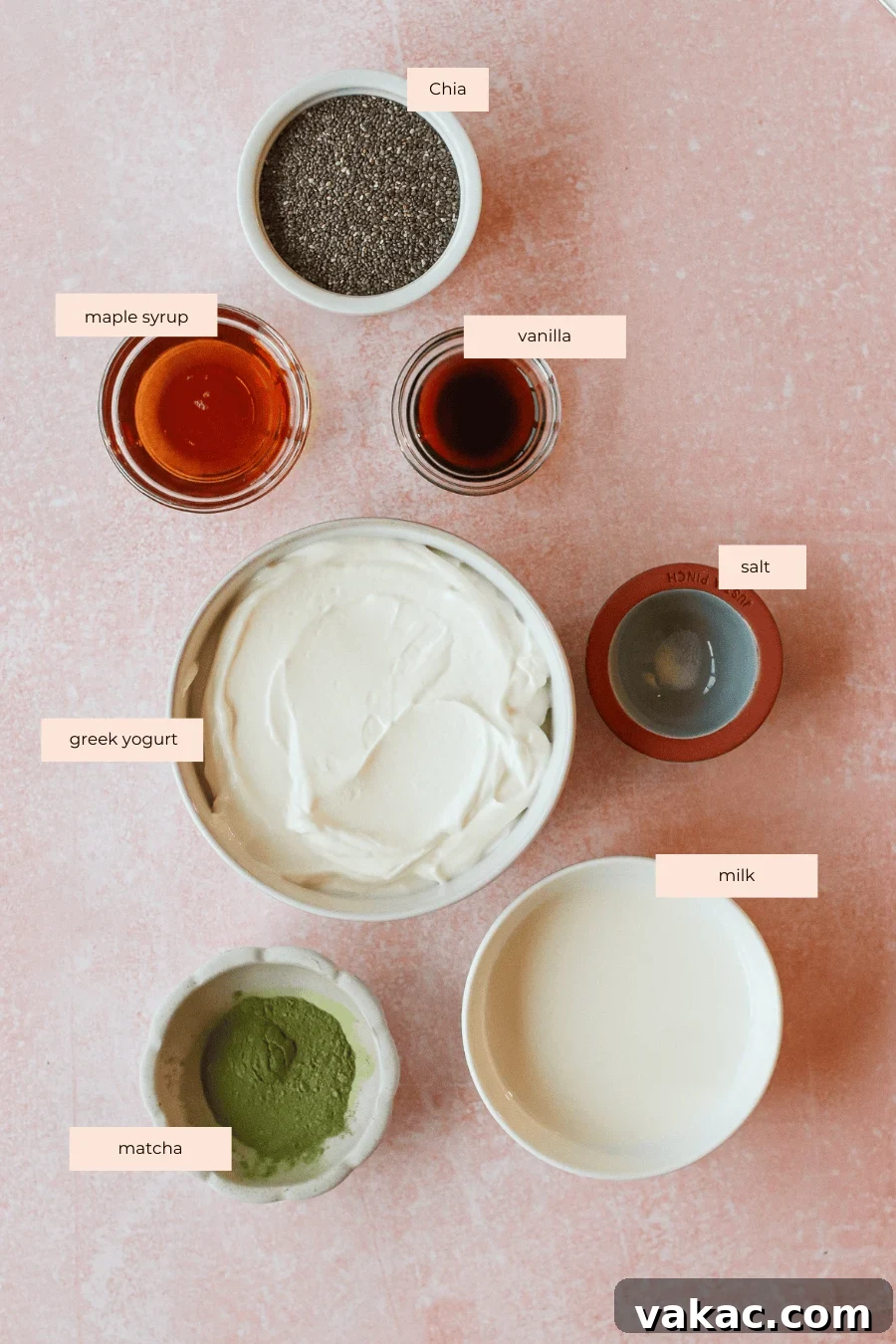 Quick High Protein Matcha Chia Pudding with Yogurt 3 A flat lay of labeled ingredients for matcha chia pudding on a pink surface: chia seeds, maple syrup, vanilla, salt, Greek yogurt, milk, and matcha powder, each in separate bowls.