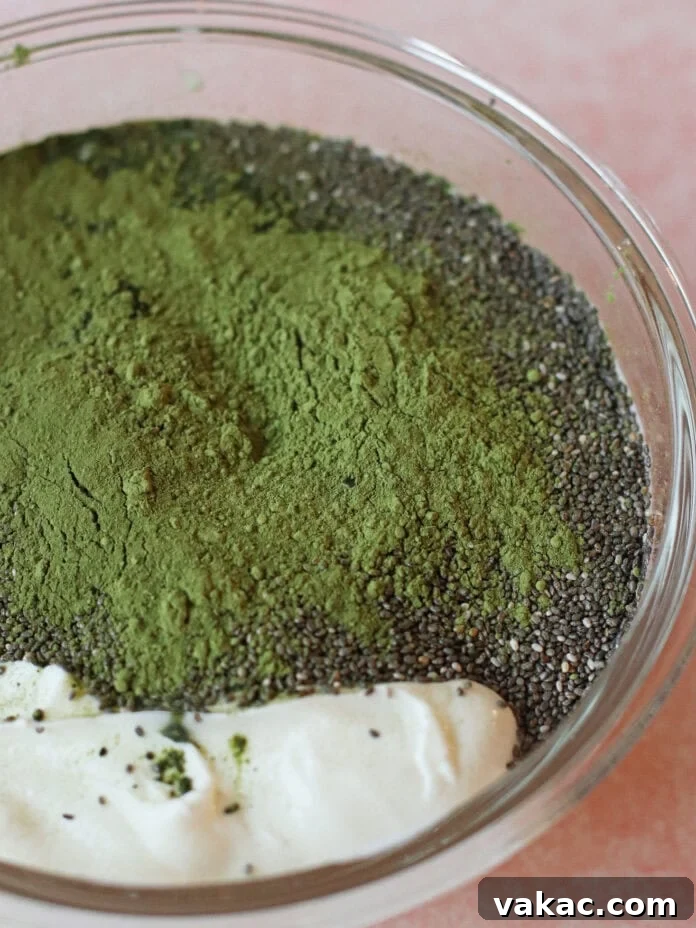 Quick High Protein Matcha Chia Pudding with Yogurt 4 A glass bowl containing chia seeds, green matcha powder, and a dollop of yogurt on a pink surface.